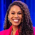 Our speaker Princess Sarah Culberson smiles during her presentation. She has long curly hair, is wearing a bright pink jacket and a headset microphone, stands in front of a blue curtain.