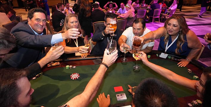 Group of adults toasting with drinks around a poker table with poker chips visible, enjoying SITE SoCal’s Poker Tournament..