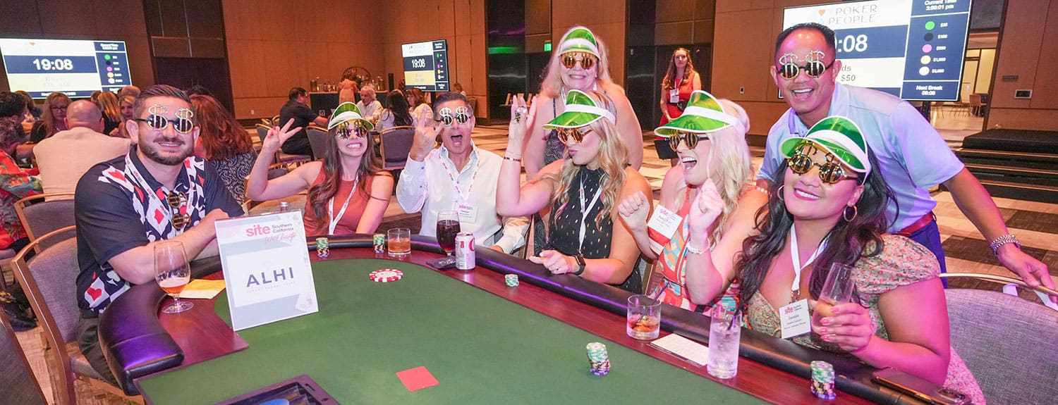 Group of people at  poker table, wearing festive hats and sunglasses, smiling and posing together at SITE SoCal’s 2023 Poker Tournament.