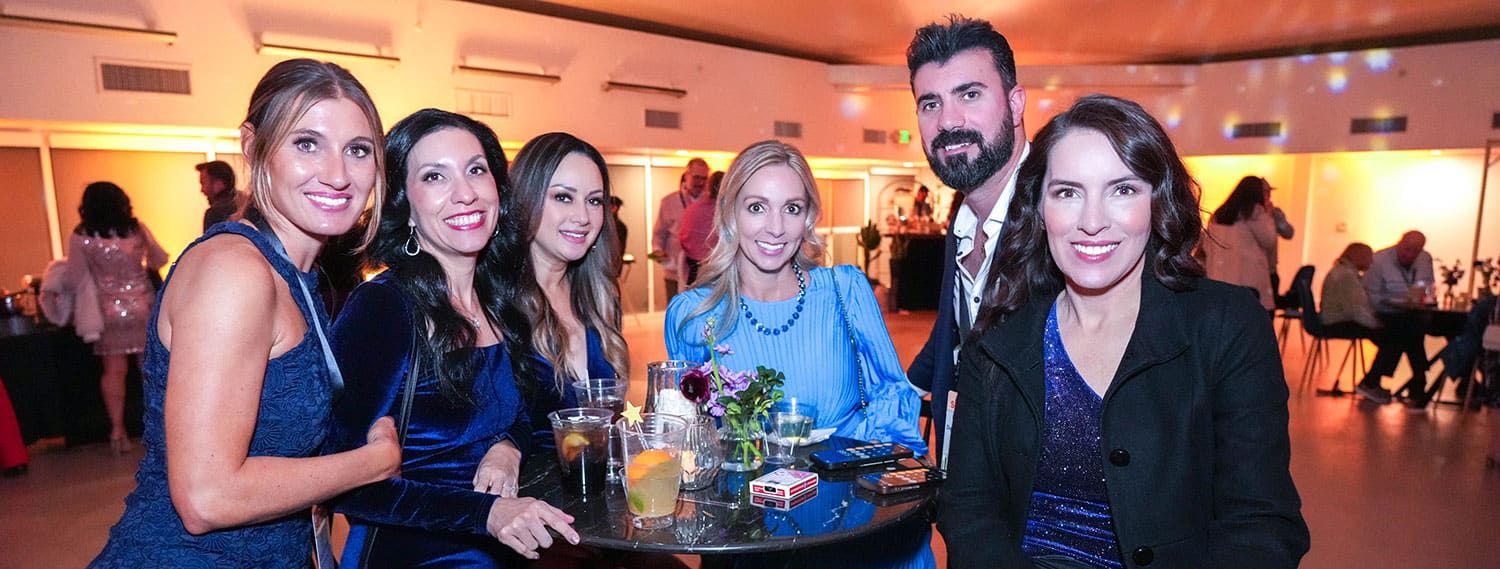 A group of people dressed in formal attire are seated and standing around a round table with drinks at SITE SoCal Holiday Event.