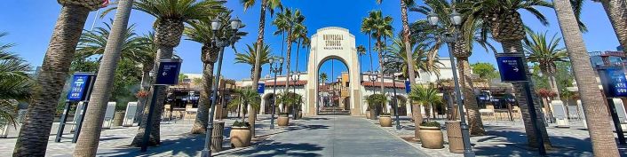 Entrance to Universal Studios Hollywood theme park featuring a prominent archway surrounded by tall palm trees, directional signs, and various buildings in the background.
