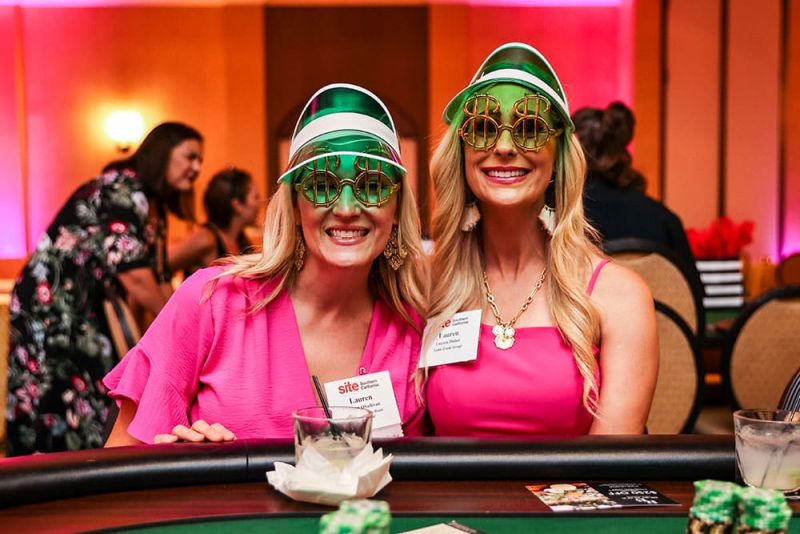 Two women wearing green visors and novelty sunglasses with dollar signs at SITE SoCal’s 2024 Poker Tournament. They are seated at a poker table, wearing bright pink outfits, and smiling at the camera. Other people are in the background.