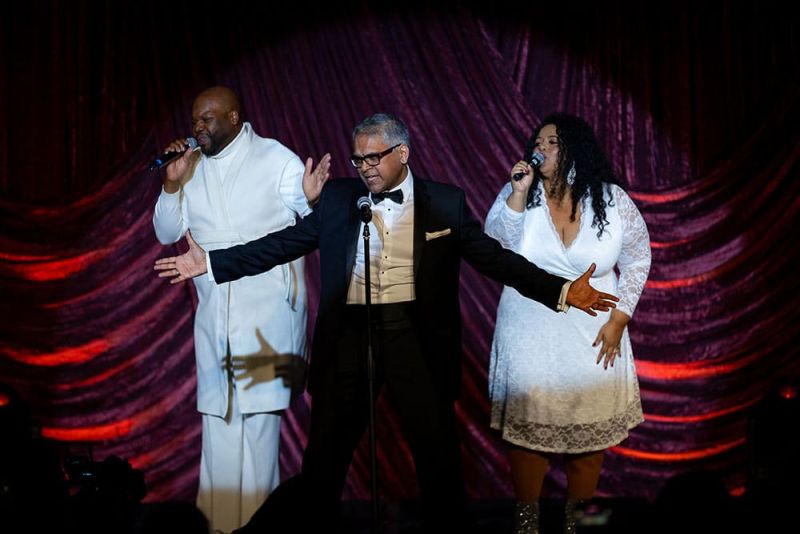 Three performers on stage sing into microphones, with Shahab Abbas in a tuxedo between two people in white outfits. A red curtain is in the background.